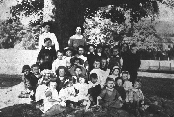 photo of schoolchildren and teachers under a tree on the green, about 1900?